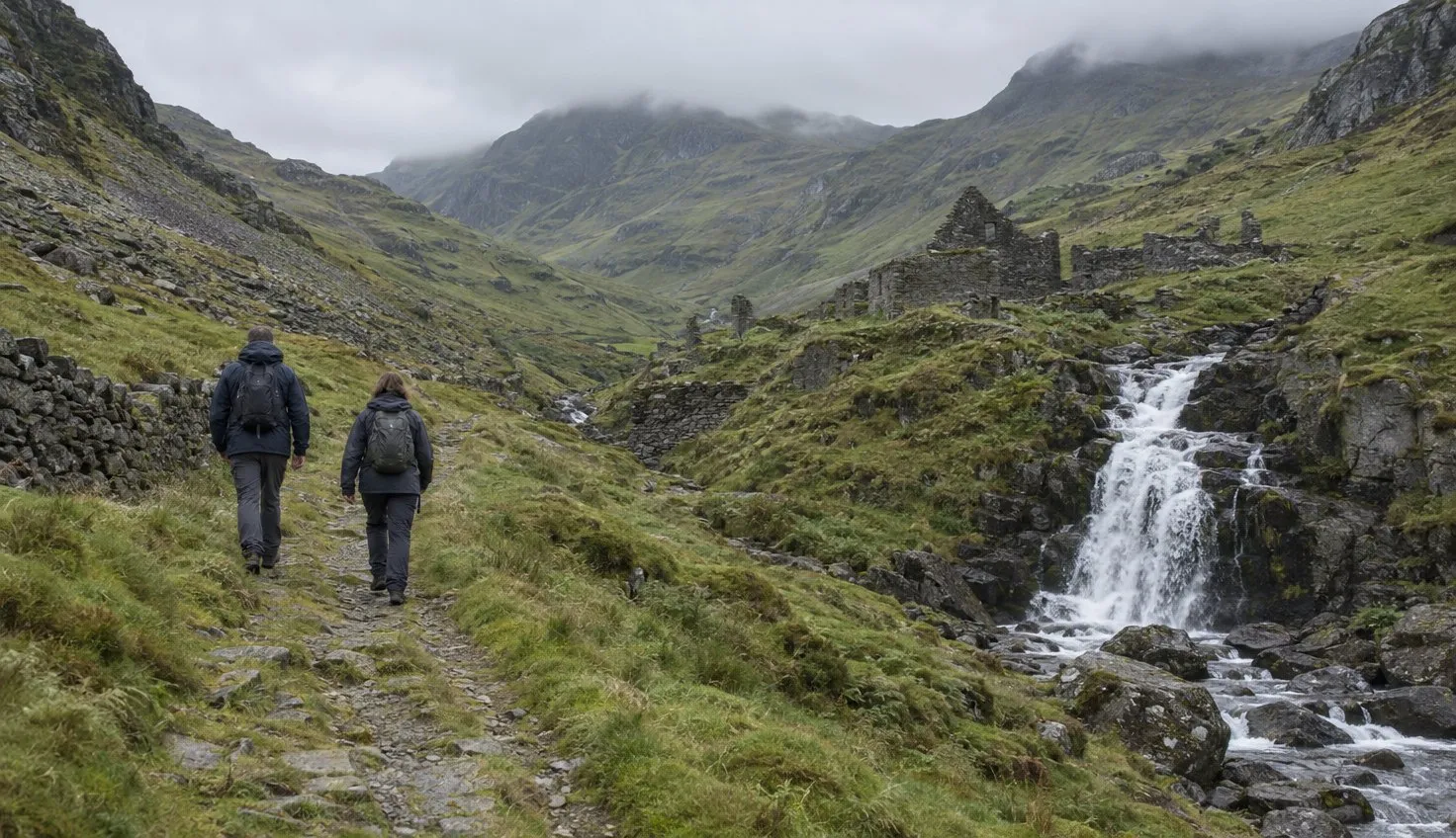 Two walkers on a grassy old track climbing into a wide mountain valley, a waterfall in the stream beside them.