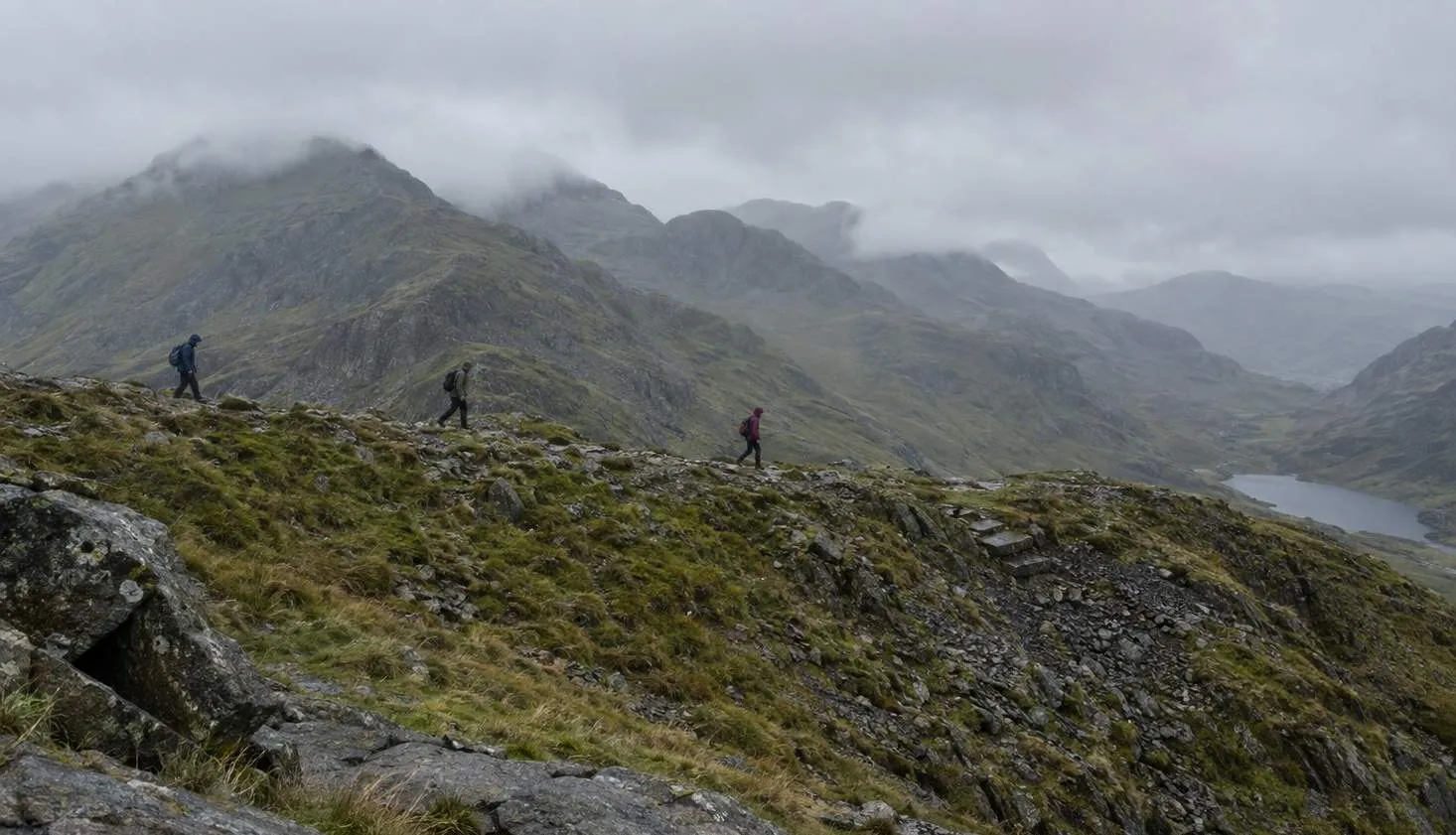 Three walkers strung out along a rocky Snowdonia ridge with more summits ahead in cloud.
