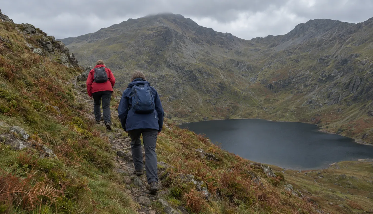 Two walkers climbing a steep grassy ridge path high above a dark corrie lake.