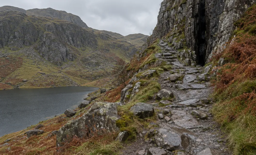 A steep rocky path climbing beside a dark cleft in a cliff above a small lake.
