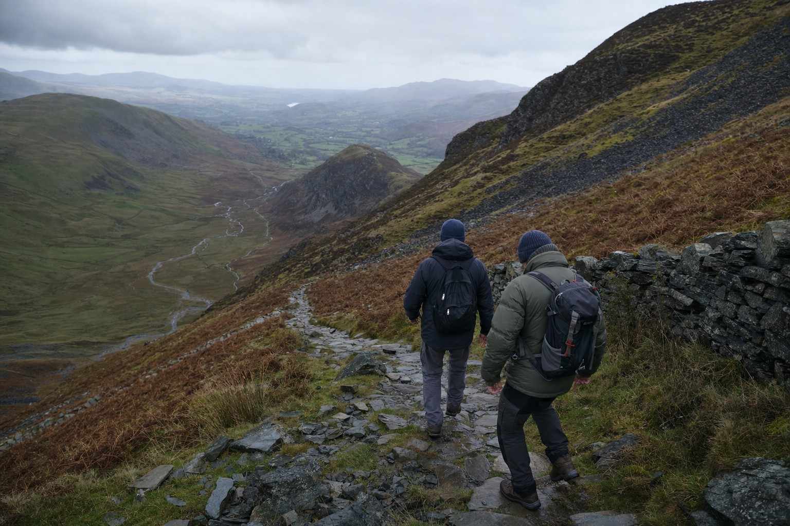 Two walkers descending a long zigzag mountain path with the valley still far below