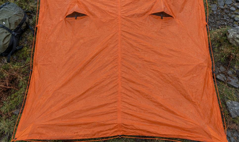 A group emergency shelter laid out flat and open on wet mountain grass, its full rectangular footprint visible from above with a walker's boots at the edge for scale.