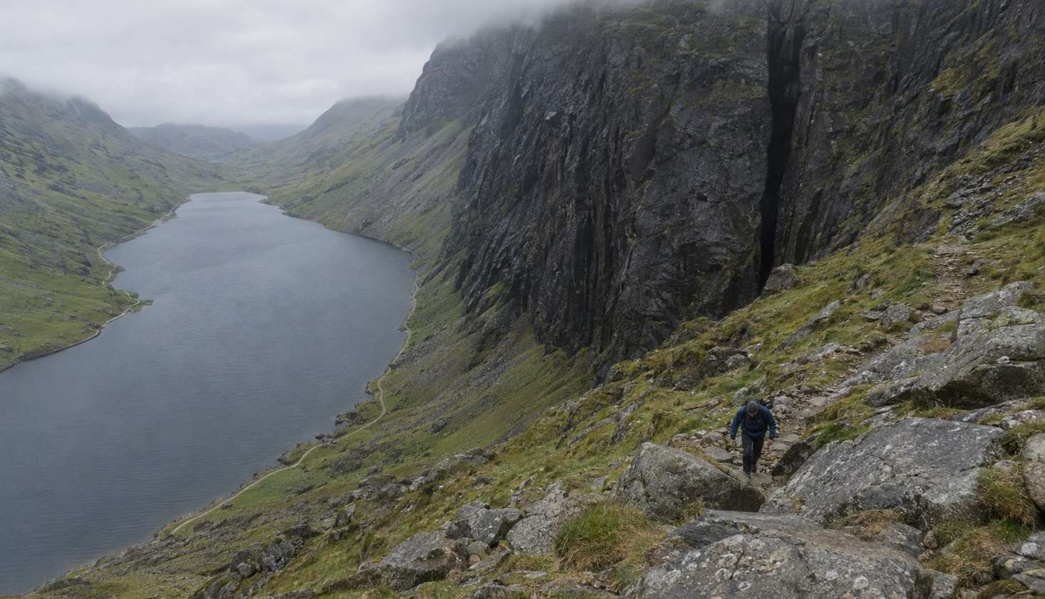 A walker on the boulder path beside the Devil's Kitchen cleft at the head of Cwm Idwal, with Llyn Idwal far below.