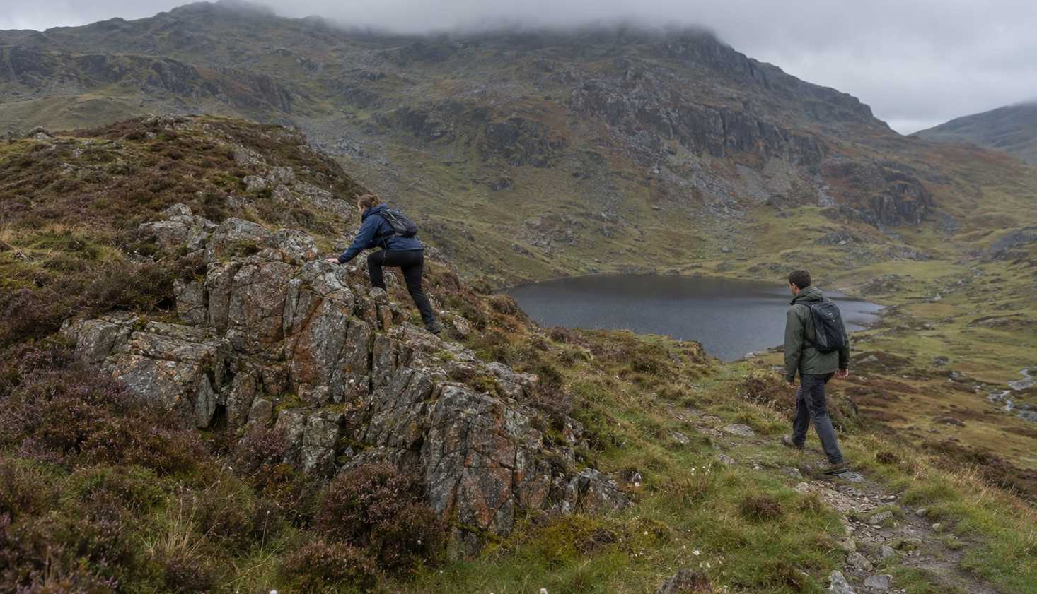 Two walkers on the Daear Ddu ridge of Moel Siabod, one scrambling on rock and one following the easier path beside it, with a small lake in the basin below.
