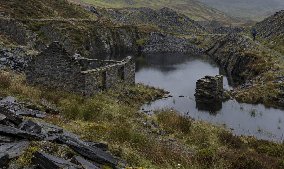 Flooded slate workings and ruined huts in the quarry basin on the way up Moel Siabod, with a walker on the path between the spoil heaps.