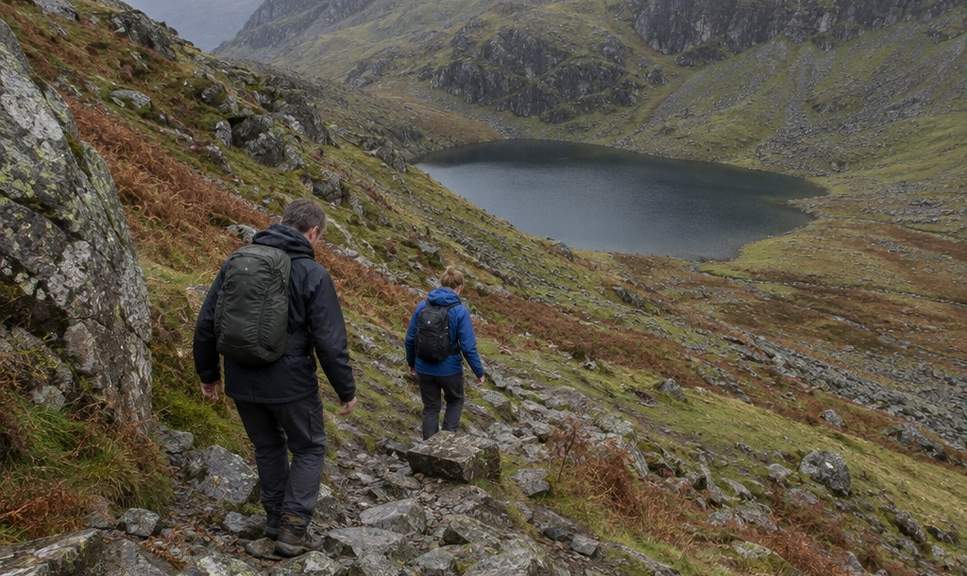 Two walkers picking their way down a rough, rocky descent below Tryfan towards a small dark mountain lake.
