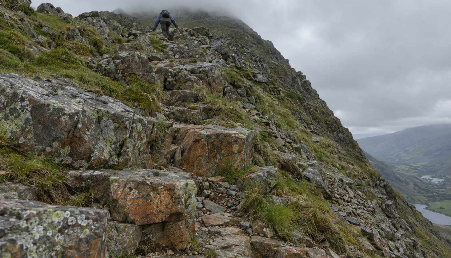 A walker using both hands on steep, rocky ground high on Tryfan's North Ridge, cloud sitting on the top and the Ogwen valley far below.