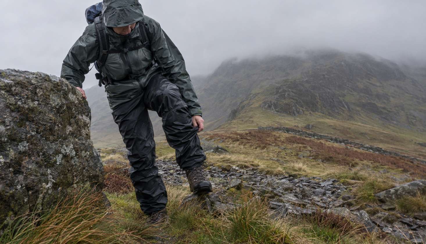 A walker on a windswept Welsh col, braced against a boulder, pulling waterproof overtrousers up over a laced walking boot while the wind pushes the fabric sideways.