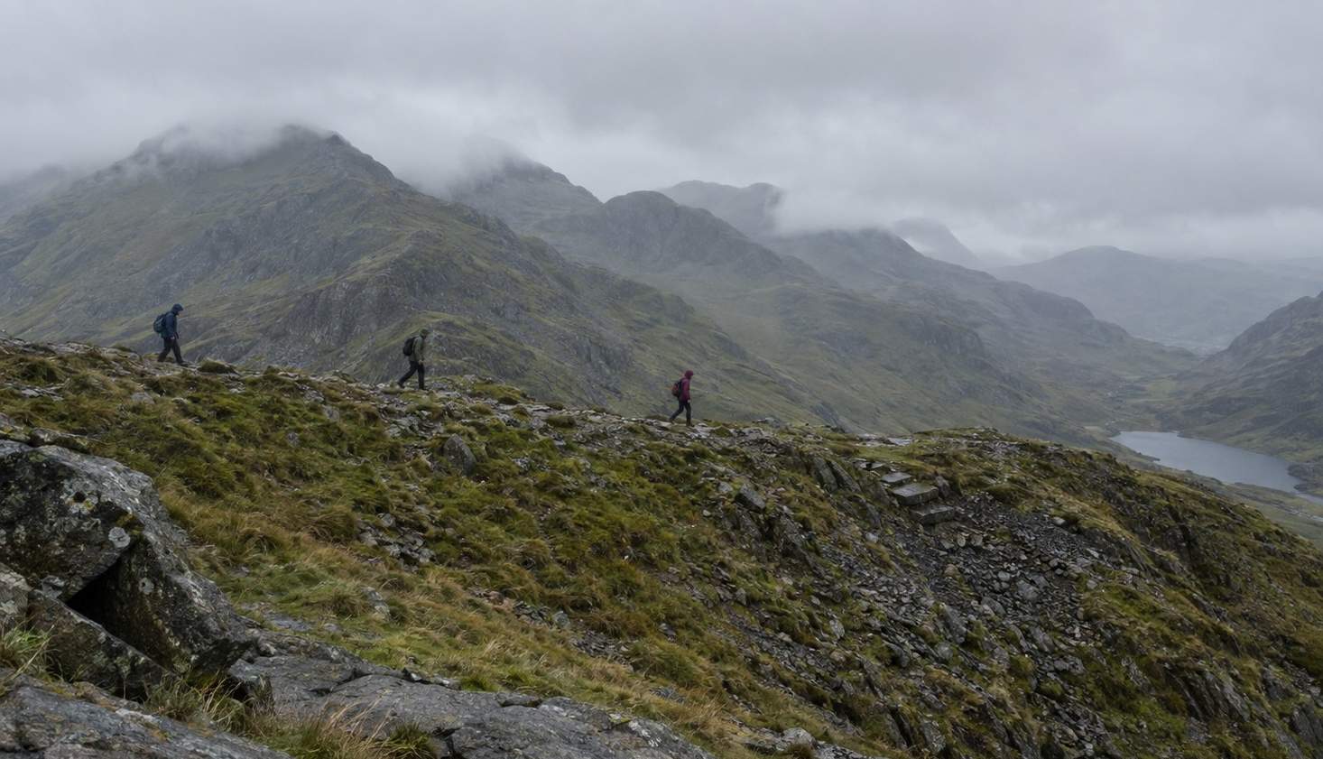 Three walkers strung out along a rocky Snowdonia ridge with more summits ahead in cloud.